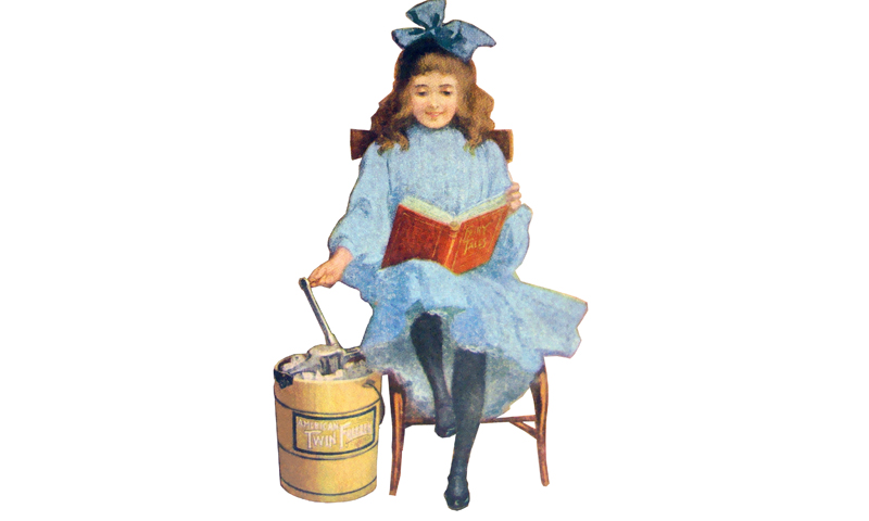 Girl in blue dress and hair bow sitting in chair reading a book while churning a hand-cranked ice cream maker.
