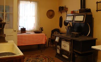 1900s kitchen with wood-burning cookstove, rag rug, and water pump at sink, and red and white gingham tablecloth.